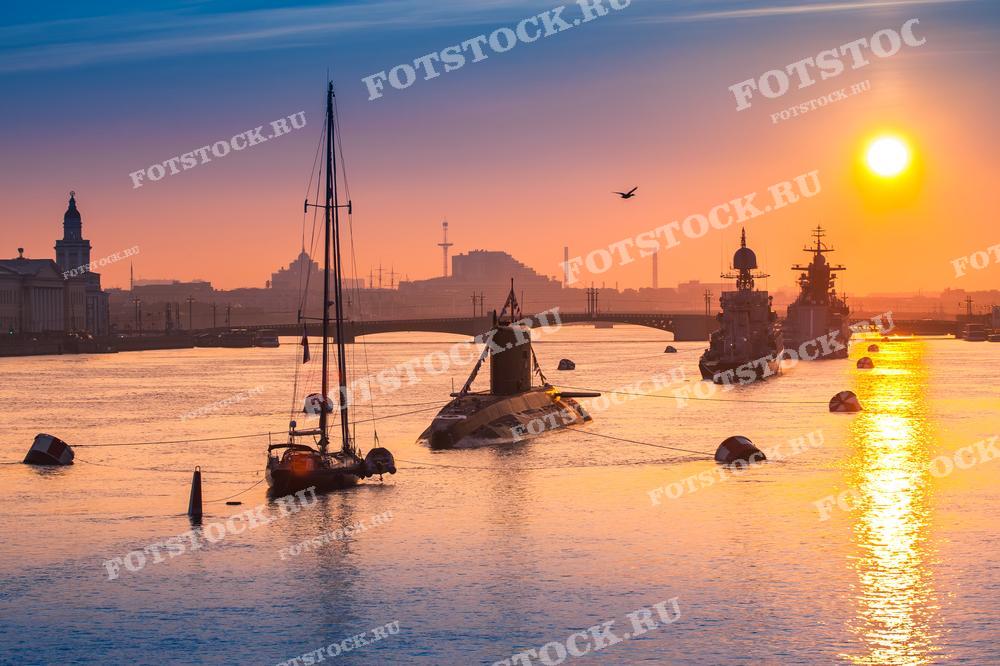 На фотографии изображены закатные лучи солнца, отражающиеся на воде реки Невы в Санкт-Петербурге. На переднем плане видны яхты и военные корабли, стоящие на якоре. Слева в кадре виднеются архитектурные сооружения Васильевского острова, а на фоне можно различить силуэты мостов и городских зданий, создающих живописный городской пейзаж.
